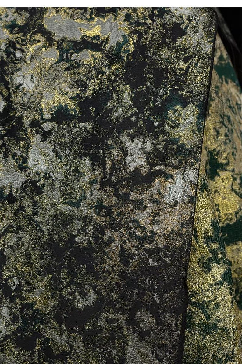 May include: Close-up of a textured fabric with a marbled design. The fabric features a mix of black, gold, silver, and green hues, creating a luxurious and abstract pattern. The material appears to be used for apparel or home decor.