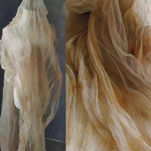 May include: A sheer fabric in a light yellow and rust brown colour. The fabric is draped and folded, creating a soft and flowing texture.