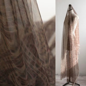 May include: A length of sheer brown fabric, patterned with a subtle design, draped over a mannequin. The fabric is semi-transparent, with a repeating pattern in brown and tan hues. The material appears lightweight and flowing, suitable for clothing or decorative use.