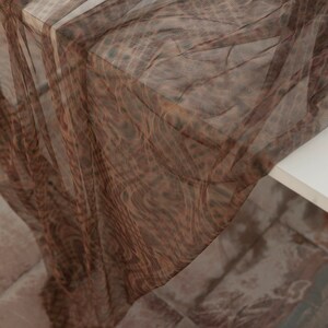 May include: A length of sheer brown fabric with a leopard print. The fabric is draped over a white surface, creating soft folds and shadows. The pattern features dark brown spots on a lighter brown background, giving it a textured appearance.