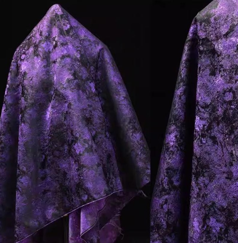 May include: A draped fabric with a marbled pattern in shades of purple and black. The fabric is folded, creating a textured surface with highlights and shadows. The material appears to be soft and flowing, suitable for apparel or home decor.