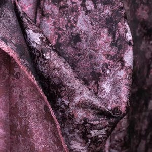 May include: Close-up of a luxurious fabric with a marbled design in shades of deep purple, black, and silver. The material has a soft, textured appearance, with visible folds and a raw edge, suggesting it's a cut piece of textile.