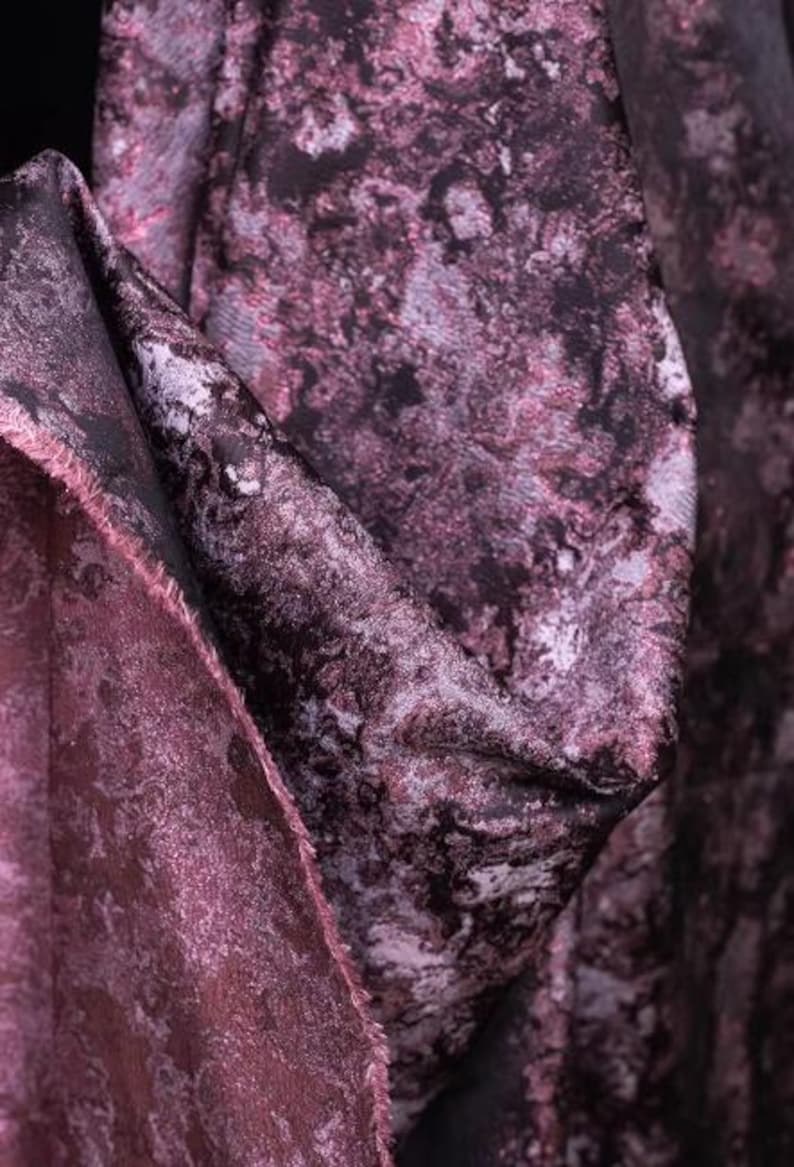 May include: Close-up of a luxurious fabric with a marbled design in shades of deep purple, black, and silver. The material has a soft, textured appearance, with visible folds and a raw edge, suggesting it's a cut piece of textile.