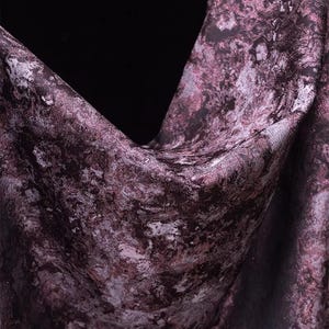 May include: Close-up of a fabric with a marbled pattern in shades of purple, gray, and black. The fabric is draped, showing folds and texture. The overall effect is a luxurious and artistic textile.