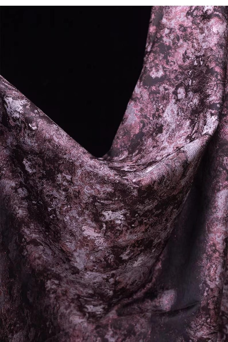 May include: Close-up of a fabric with a marbled pattern in shades of purple, gray, and black. The fabric is draped, showing folds and texture. The overall effect is a luxurious and artistic textile.