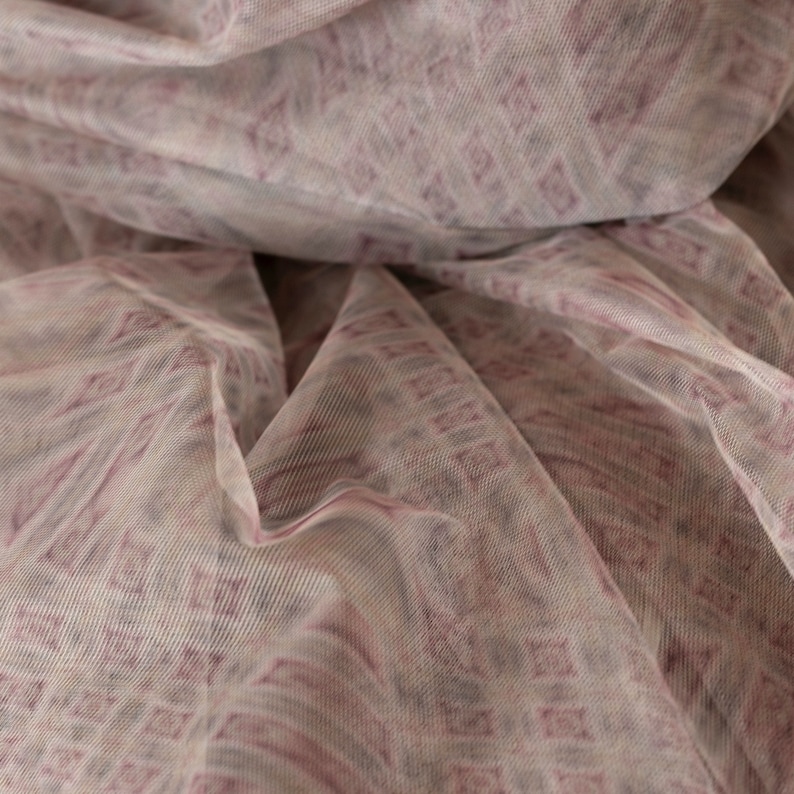 May include: Close-up of a sheer fabric with a repeating diamond pattern in shades of mauve and off-white. The fabric is draped, creating soft folds and shadows. The material appears lightweight and delicate, suitable for apparel or decorative purposes.
