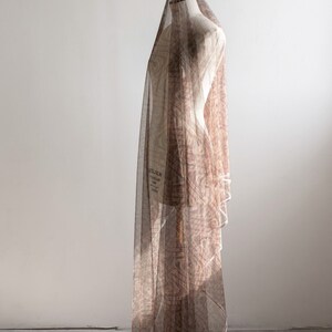 May include: A dressmaker's mannequin covered with a patterned, sheer fabric. The fabric is a light brown colour and drapes over the form. The mannequin is on a black metal stand, against a white wall.