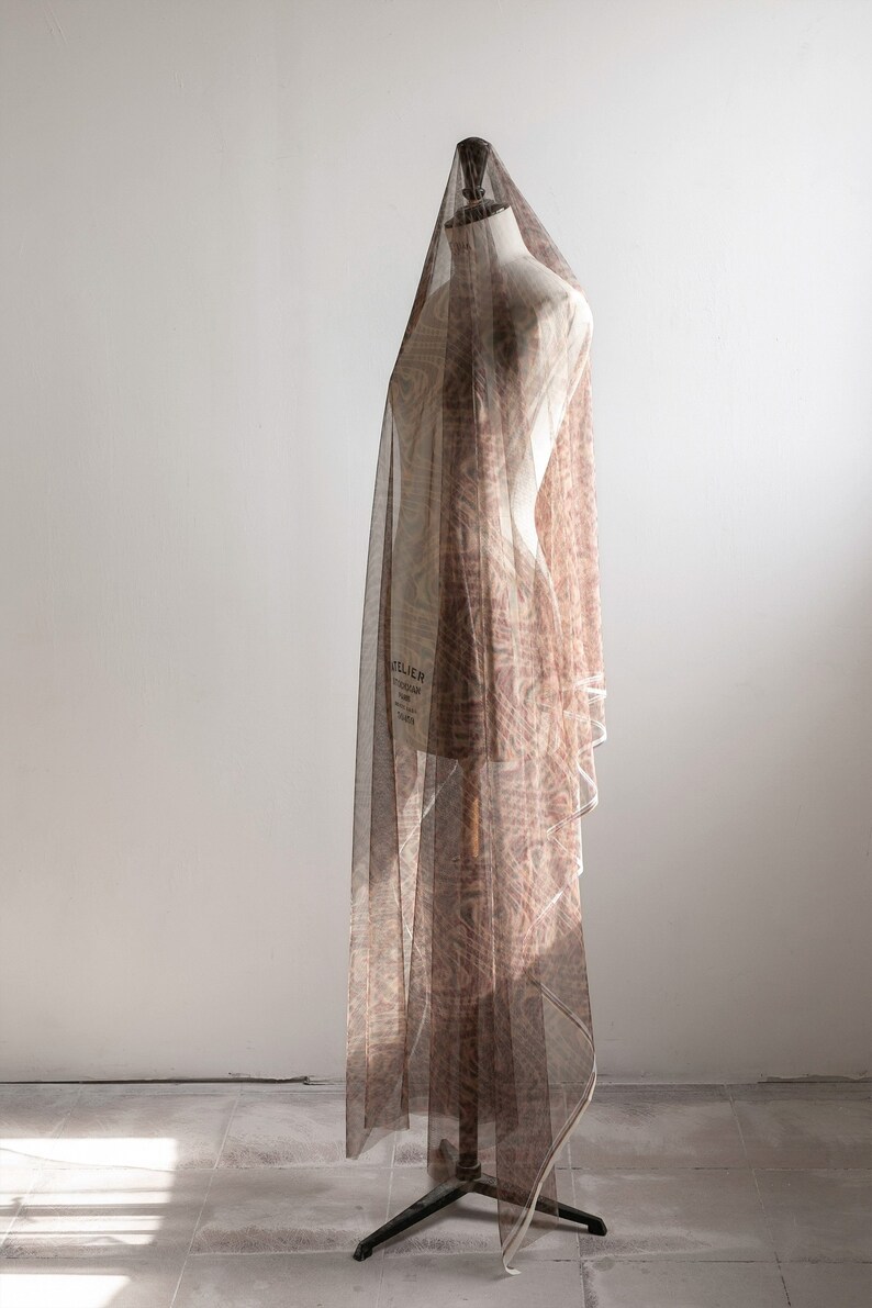 May include: A dressmaker's mannequin covered with a patterned, sheer fabric. The fabric is a light brown colour and drapes over the form. The mannequin is on a black metal stand, against a white wall.