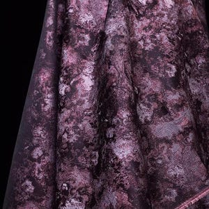 May include: Close-up of a draped fabric with a marbled pattern in shades of deep purple, lavender, and silver. The fabric has a textured appearance and a subtle sheen, with visible folds and a raw edge.
