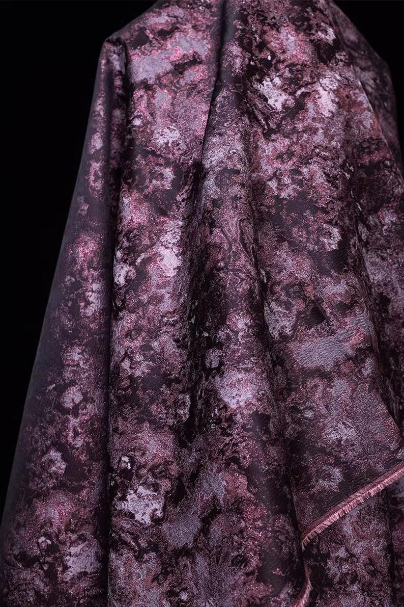 May include: Close-up of a draped fabric with a marbled pattern in shades of deep purple, lavender, and silver. The fabric has a textured appearance and a subtle sheen, with visible folds and a raw edge.