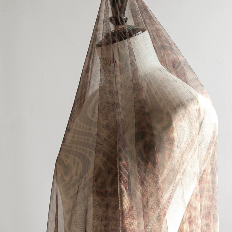 May include: Sheer brown fabric draped over a mannequin. The fabric features a patterned design with shades of brown and red. The fabric is pleated, creating vertical stripes. The mannequin is a neutral colour, providing a contrast to the fabric.