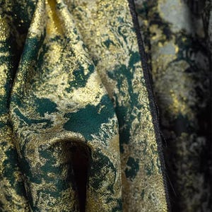May include: Close-up of a luxurious fabric with a shimmering gold and deep emerald green pattern. The textile has a textured appearance, with the colors blending to create a rich, opulent look. The fabric's folds add depth.