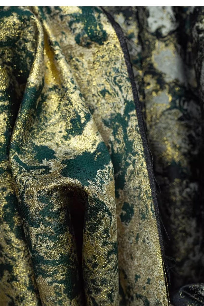 May include: Close-up of a luxurious fabric with a shimmering gold and deep emerald green pattern. The textile has a textured appearance, with the colors blending to create a rich, opulent look. The fabric's folds add depth.