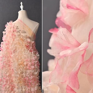 May include: A one-shoulder dress featuring a sheer base fabric adorned with numerous layered, translucent fabric petals in shades of white and soft pink. The petals create a dense, textured floral effect across the entire surface of the garment.
