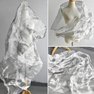 May include: A sheer, silver-toned organza fabric draped over a mannequin. The material is translucent with a metallic sheen, featuring a crinkled, lightweight texture that creates voluminous folds and ripples across the form of the display.