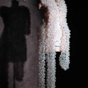 Puede incluir: Una prenda rosa claro, con textura de burbujas, drapeada sobre un maniquí. La prenda parece ser una bufanda o chal, con largos mechones colgantes. El fondo es oscuro, resaltando la textura y la forma de la pieza.