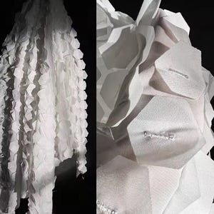 May include: White fabric with a geometric design. The material is layered, creating a 3D effect. The intricate design highlights the interplay of light and shadow. The overall aesthetic is modern and architectural.