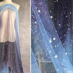 Op de afbeelding: Een blauwe en paarse tule stof met een gradiënt, witte glitter en parelversieringen. De stof is doorschijnend en heeft een zachte, vloeiende textuur.