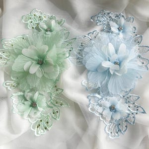 May include: Two floral appliques, one in light green and the other in light blue, each featuring layered fabric petals and sparkling rhinestone centers. Delicate lace and beaded accents surround the flowers, creating an elegant, decorative design.