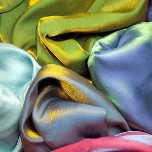 Könnte beinhalten: Nahaufnahme eines Stapels schimmernder Stoffe in Grün-, Blau- und Rosatönen. Der Stoff hat einen dezenten Schimmer und ist in einem weichen, fließenden Muster angeordnet.