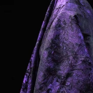 May include: A close-up of a draped fabric with a textured, marbled appearance. The fabric is primarily a deep purple color with black accents, creating a dramatic and luxurious look. The folds and shadows add depth.