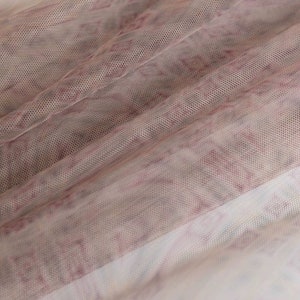 May include: Close-up of a sheer fabric with a delicate pattern. The fabric is a light, airy material with a mesh-like texture. The pattern features a mix of pink and white hues, creating a soft, textured look. The fabric is draped, showing folds and shadows.