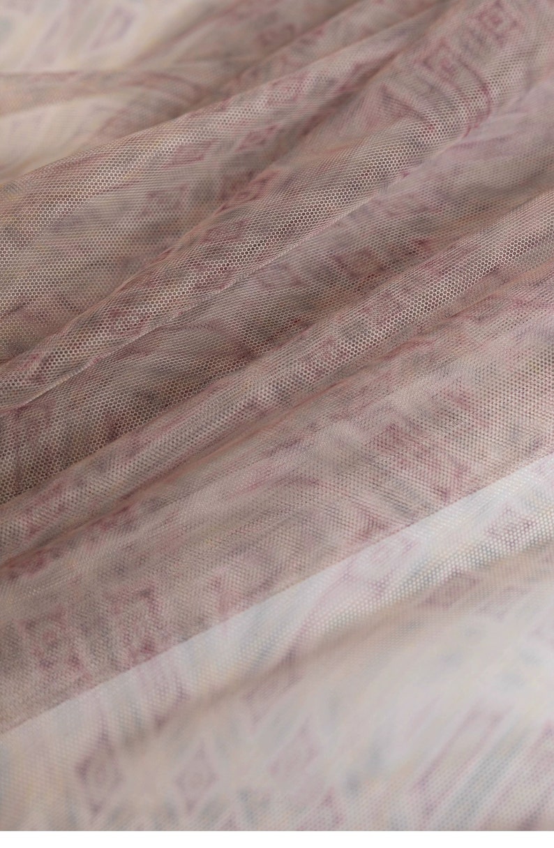May include: Close-up of a sheer fabric with a delicate pattern. The fabric is a light, airy material with a mesh-like texture. The pattern features a mix of pink and white hues, creating a soft, textured look. The fabric is draped, showing folds and shadows.