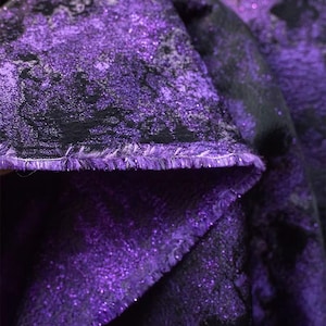 May include: Close-up of a folded fabric with a shimmering purple and black pattern. The fabric has a textured surface with a glittery effect, and the edges are frayed. The overall theme is textile and design.