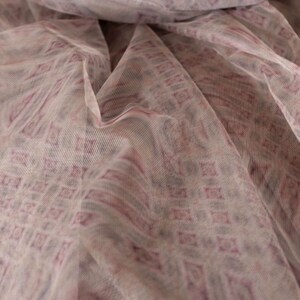 May include: Close-up of a sheer fabric with a repeating geometric pattern. The fabric is a light, neutral colour with a pattern in shades of mauve and purple. The fabric is draped, creating soft folds and shadows.