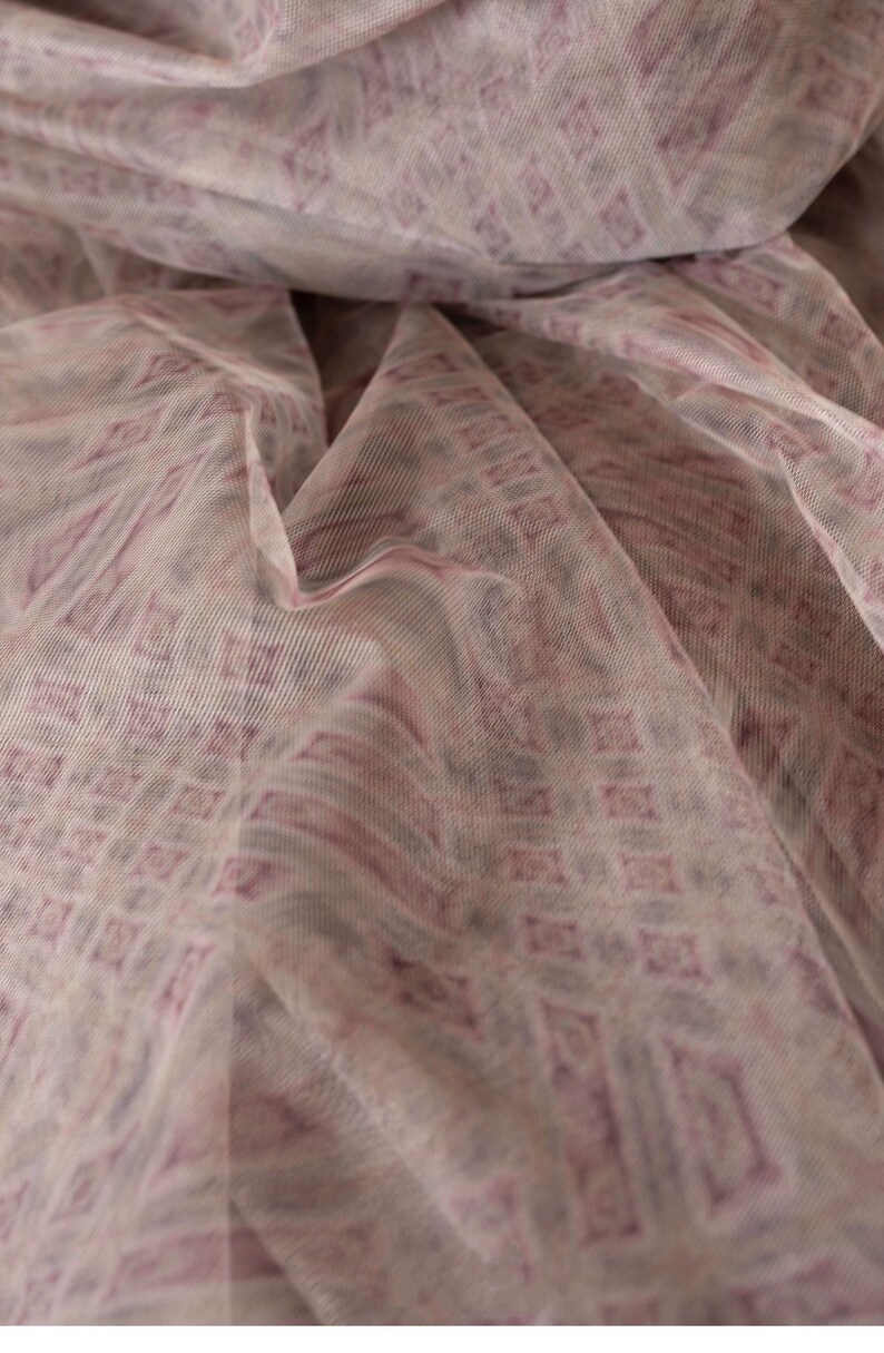 May include: Close-up of a sheer fabric with a repeating geometric pattern. The fabric is a light, neutral colour with a pattern in shades of mauve and purple. The fabric is draped, creating soft folds and shadows.