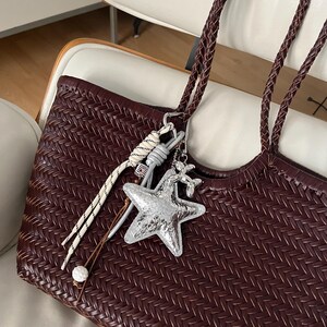Peut inclure: Un sac fourre-tout bordeaux tissé avec des anses tressées. Le sac est orné d'un charm en forme d'étoile argentée et d'autres breloques décoratives. Le sac est posé sur une chaise blanche.