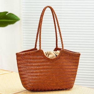 May include: A woven brown tote bag with braided handles. The bag has a rectangular shape with a curved opening and a beige fabric lining visible inside. The bag is sitting on a light-colored wooden surface.