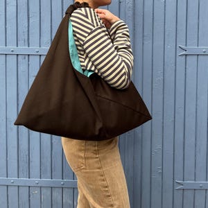 Peut inclure: Un sac fourre-tout marron foncé de forme triangulaire. Le sac a un intérieur bleu clair et une fermeture à cordon. La personne porte une chemise à manches longues rayée et un pantalon beige. L'arrière-plan est une porte en bois bleue.