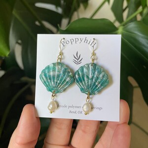 May include: A pair of handmade polymer clay earrings in a seashell design. The earrings are teal with gold accents and feature a freshwater pearl drop. The earrings are on a white card with the text "poppyhill" and "Bend, OR".