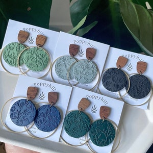 May include: Four pairs of handmade polymer clay earrings with a leafy design. Each pair features a different color of green, blue, and black. The earrings are attached to gold hoops and have wooden tops.