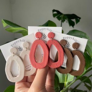 May include: Three pairs of polymer clay earrings in different colors. The earrings are shaped like an oval with a cutout in the center. The earrings are textured with a leaf pattern. The earrings are handmade by Poppyhill.
