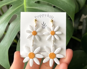 May include: White polymer clay daisy earrings with a yellow center. The earrings are on a white card with the text "Poppyhill polymer" printed on it.