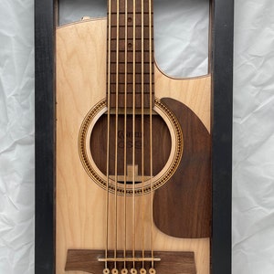 May include: A framed wooden cutout of an acoustic guitar with a black frame. The guitar is made of light and dark wood and has a sound hole with a decorative inlay. The guitar has six strings and a bridge with six tuning pegs.