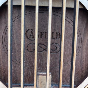 May include: Close-up of the soundhole of an acoustic guitar with a wooden soundboard. The soundhole is surrounded by a decorative inlay. The brand name "Canfield" is engraved in the wood.