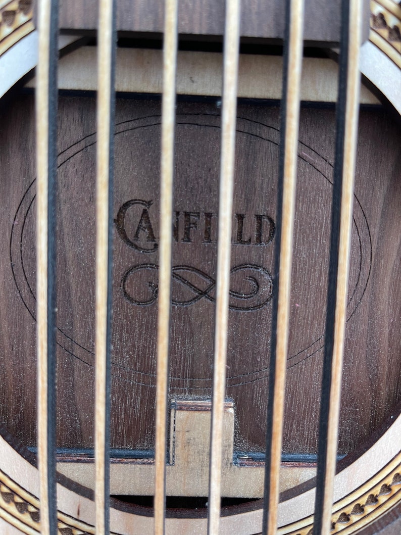 May include: Close-up of the soundhole of an acoustic guitar with a wooden soundboard. The soundhole is surrounded by a decorative inlay. The brand name "Canfield" is engraved in the wood.