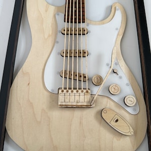 May include: A wooden model of an electric guitar in a black frame. The guitar is made of light wood and has a white pickguard. The guitar has six strings, a bridge, and a volume and tone knob.