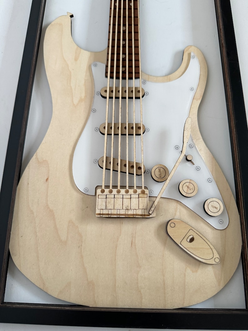 May include: A wooden model of an electric guitar in a black frame. The guitar is made of light wood and has a white pickguard. The guitar has six strings, a bridge, and a volume and tone knob.