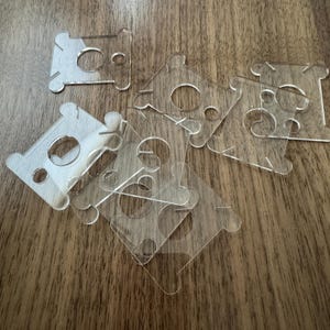May include: A collection of clear, acrylic thread spool holders scattered on a wooden surface. Each holder has a central circular opening and four smaller holes, with a unique shape designed to secure thread spools. The transparent material allows for easy visibility.