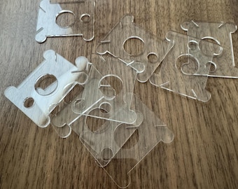 May include: A collection of clear, acrylic thread spool holders scattered on a wooden surface. Each holder has a central circular opening and four smaller holes, with a unique shape designed to secure thread spools. The transparent material allows for easy visibility.