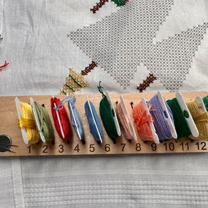 Puede incluir: Un organizador de hilos de madera con 12 ranuras numeradas, cada una con un color diferente de hilo de bordar. Los colores de los hilos son amarillo, verde, rojo, azul, verde, beige, naranja, morado, verde, amarillo y blanco.