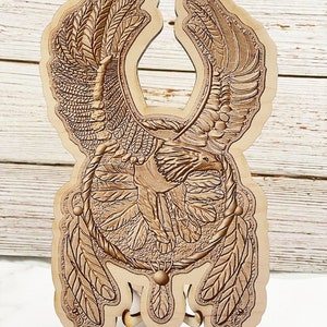 May include: A wooden decorative piece featuring an eagle with outstretched wings, surrounded by feathers and a circular design. The piece is carved with intricate details and has a natural wood finish.