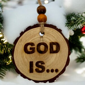 Puede incluir: Un adorno navideño de madera con el texto "GOD IS..." El adorno está colgado de una cuerda con cuentas marrones y naranjas. El adorno está sobre una superficie nevada.