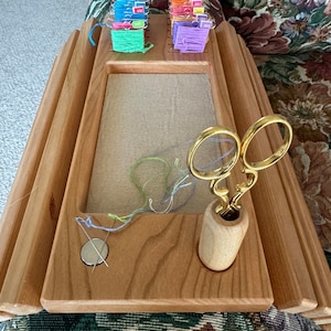 May include: Wooden embroidery hoop stand with a built-in thread organizer and a compartment for scissors. The stand has a light wood finish and is sitting on a floral patterned fabric.