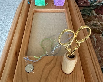 May include: Wooden embroidery hoop stand with a built-in thread organizer and a compartment for scissors. The stand has a light wood finish and is sitting on a floral patterned fabric.