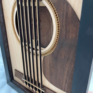 May include: A close-up of a wooden acoustic guitar with a black frame. The guitar has a sound hole with a decorative border and a wooden bridge with six tuning pegs.